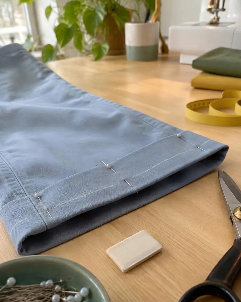 Blaue Stoffhose mit gestecktem Saum auf dem Werktisch, daneben Schere und Schneiderkreide